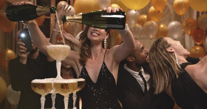 Woman pouring champagne into tower of glasses with friends dancing around and shooting a film on their mobile phone at gala night party.
