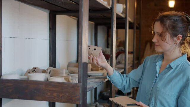 Slow motion of serious young woman looking at hand-made earthware then taking notes working alone in workshop. Pottery business and people concept.