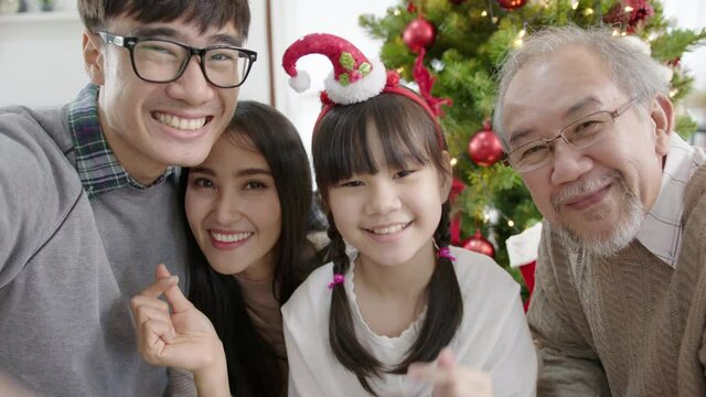 Point of view mobile screen of happy asian family in quarantine merry christmas enjoy selfie or take photo shoot group shot sitting together at home apartment enjoy leisure time with X'mas tree.