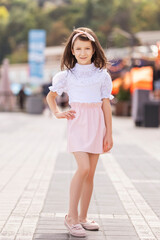 Little brunette girl in blouse and skirt posing alone on european street on summer day.
