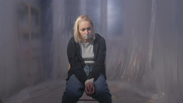Female hostage sitting on a chair in a room with her mouth taped shut