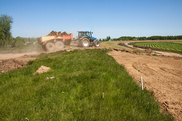 Fototapeta premium Tractor with tipper trailer loaded with clay drives down a dike on a dusty track; in the background a green polder and a reenforced dike with a new layer of clay. 