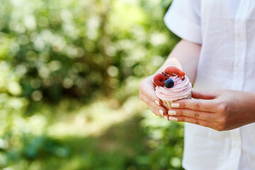 Appetizing cupcake covered with pink cream and summer berries in the woman's hands. Green bokeh background, focus on the right side. Copy space. 
