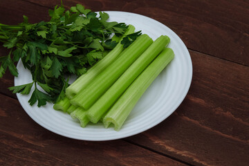 Green sliced celery stalks and a bunch of parsley on a white plate close-up and on a brown wooden background-the concept of cooking healthy food, salads and soups, weight loss, slimming and  supper