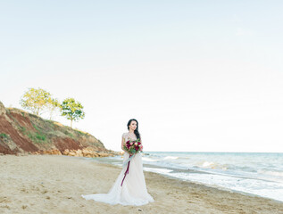 Beautiful brunette bride in light chiffon wedding dress embroidered with beads posing near the sea. Romantic beautiful bride in luxury dress posing on the beach.