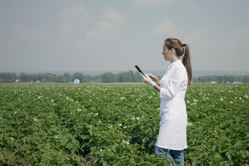 Young female agronomist in white lab coat using digital tablet pc while working in agricultural field. Concept ecology, bio product, inspection, natural products, professional