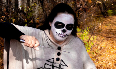 woman with creative Halloween skull in autumn forest with a large knife in her hands. A woman prepares for the Mexican Day of the Dead. Horror beauty nightmares.