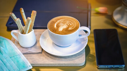 Hot Cappuccino coffee with protective mask on a wooden table for people being quarantined in the situation of the spread of the corona virus (Covid-19).  New normal lifestyle concept. Selective focus