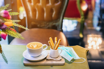 Hot Cappuccino coffee with protective mask on a wooden table for people being quarantined in the situation of the spread of the corona virus (Covid-19).  New normal lifestyle concept. Selective focus