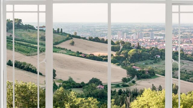 Bolognese hills landscape as seen from a white window while birds are flying at the sunset. It is seen wheat fields and trees in close up and Bologna city in background
