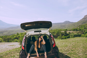 Young beautiful woman in a car trunk of a hatchback on a background of a picturesque mountain landscape.