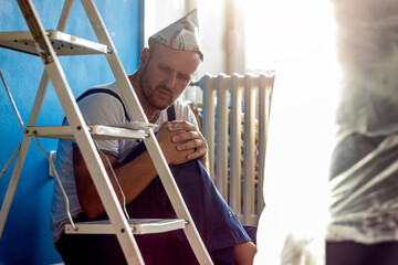 Craftsman sitting next to the ladder with pain in the knee. Knee injury at work of painter worker at home. Man worker with knee injury, concept of accident at work. Healthcare and medical concept.