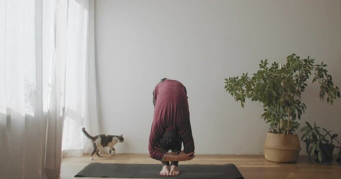 Fit man standing in uttanasana pose on mat indoors natural light slow motion. Yogi master demonstrating flexible body practicing yoga in sunny room copy text space. Active healthy lifestyle concept