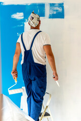 Rear view of a young man painting wall at home in white paint. Man standing on ladder while decorating domestic room with paint roller in hand. Portrait of painter man holding paint roller and brush.