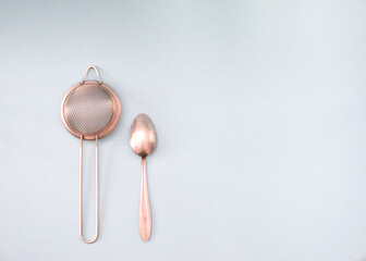 A metal tea strainer next to a stainless steel teaspoon isolated on a light background.