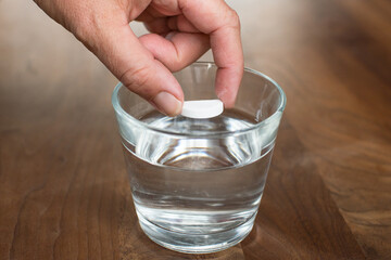 Woman hand putting fizzy tablet to the glass of water on wooden table. Vitamin and magnesium tablet in drink.