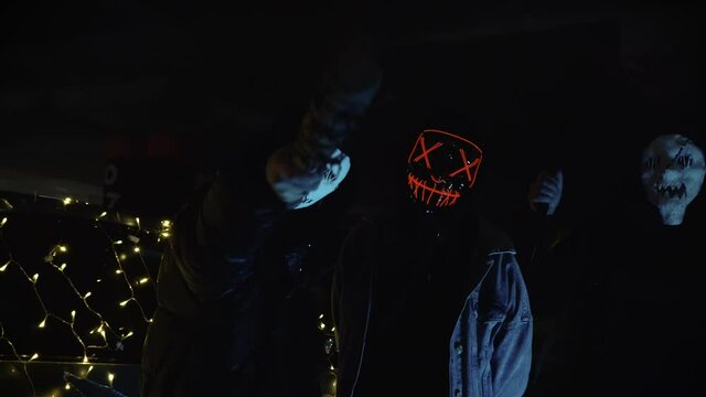 People with luminous purge masks are holding a baseball bat and katana. Murderers at night in the parking lot against the backdrop of a car with a garland.