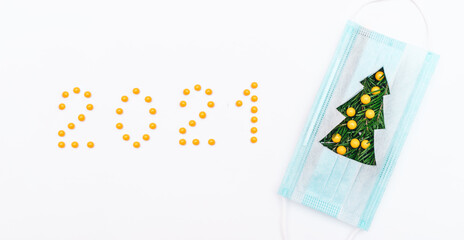 Medical protective mask with a branches of fir tree on a white background.Figures 2021 and Christmas toys laid out with yellow vitamin pills.Christmas and Happy New Year concept, medical concept.
