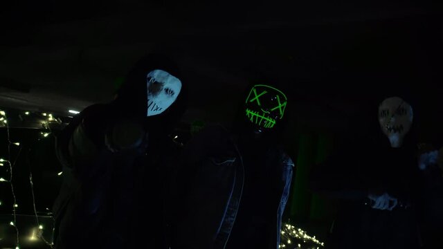 People with luminous purge masks are holding a baseball bat and katana. Murderers at night in the parking lot against the backdrop of a car with a garland.