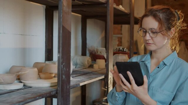 Modern young woman is working with tablet in pottery workshop touching screen looking at ceramics busy with inventory. People and technology concept.