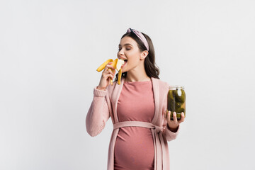 joyful pregnant woman eating banana and holding jar with canned cucumbers isolated on white