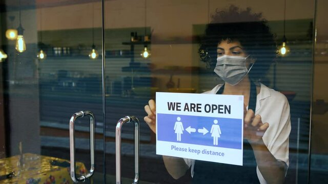 Female owner wearing surgical mask, reopens after lockdown quarantine. African american businesswoman hangs an open sign, after Coronavirus outbreak. New normal, keep the distance concept.