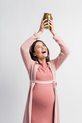 excited and pregnant woman holding jar with pickled cucumbers above head isolated on white