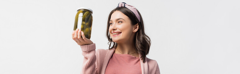 panoramic shot of brunette woman in headband looking at jar with pickled cucumbers isolated on white