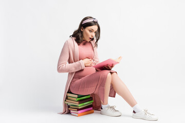 shocked and pregnant woman sitting on books and touching belly on white