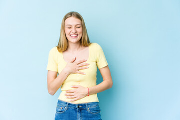 Young blonde woman isolated on blue background laughs happily and has fun keeping hands on stomach.