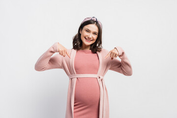 joyful and pregnant woman pointing with fingers at belly on white