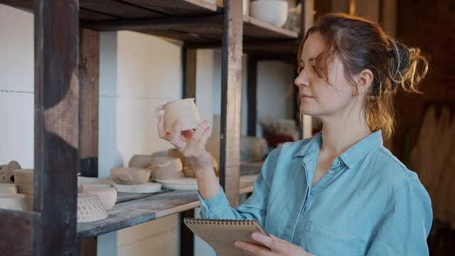 Slow motion of serious female potter checking ceramics and writing notes in workshop concentrated on family business. People and inventory concept.