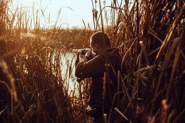 Young hunter man with a shotgun hiding in the reeds near the pond, at the duck hunt - photo with...