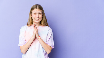 Young blonde woman isolated on purple background holding hands in pray near mouth, feels confident.