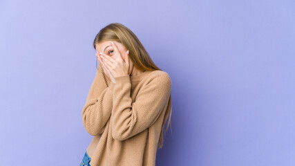 Young blonde woman isolated on purple background blink through fingers frightened and nervous.