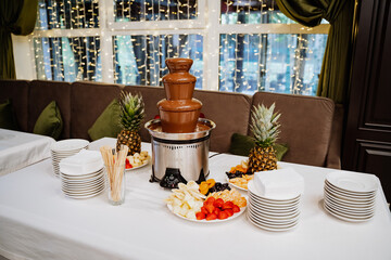 a chocolate fountain stands on a white table with sliced fruit next to it. dessert table. Sweet...