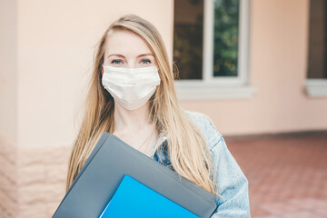 Female student wearing a medical protective respiratory mask holds notebook folders in her hands and looks at the camera