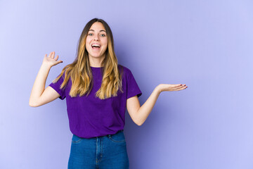 Young caucasian woman isolated on purple background holds copy space on a palm, keep hand over cheek. Amazed and delighted.