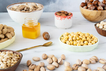 An assortment of nuts (hazelnuts, almonds, cashews, pine nuts) and chickpeas in different bowls are on a white table. There is a jar of honey in the center. Below are scattered pistachio nuts in shell