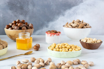 Pistachios and different nuts (hazelnuts, almonds and cashews) in different wooden and ceramic bowls stand on a gray-white background. In-shell pistachios sprinkled along the bottom edge