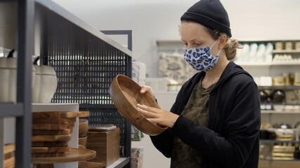 Young woman wearing protective face mask chooses wooden pottery in in dinneware department of home decor shop