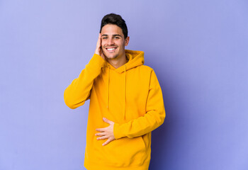 Young caucasian man isolated on purple background laughs happily and has fun keeping hands on stomach.