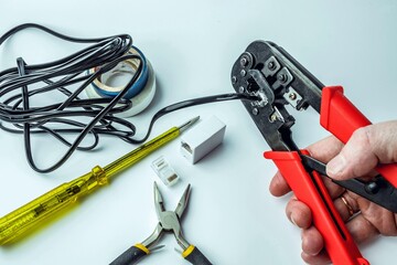 Hand of a craftsman with a crimping tool connecting an RJ45 connector with a black cable on a white background with a screwdriver and pliers.