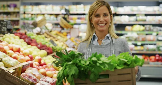 Authentic shot of friendly female staff worker wearing apron is carrying a wooden crate with the fresh vegetables is smiling in camera satisfied with her work in organic department at supermarket