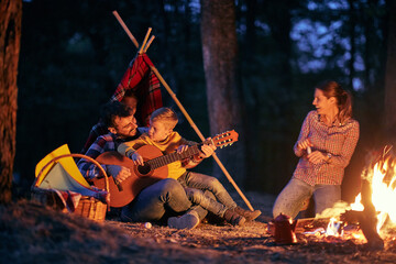 A young happy family enjoying a guitar and a campfire in the forest