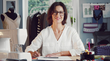 Smiling female dressmaker sitting at table in front of sheet of paper working in sewing studio