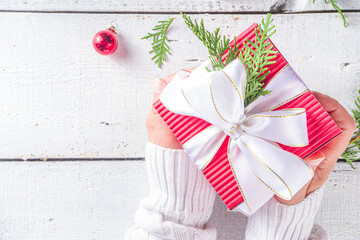 Female hands hold gift box on white wooden background, copy space. Girl in white sweater holds gift box in red wrapping paper with white festive ribbon. Christmas, New year background