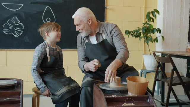 Caring senior grandfather is showing grandson how to work with clay on throwing-wheel in small workshop. Pottery, family hobby and handicraft concept.