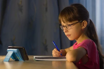 Deurstickers Online onderwijs op afstand. Schattig schoolmeisje kijkt naar mobiel © Albert Ziganshin
