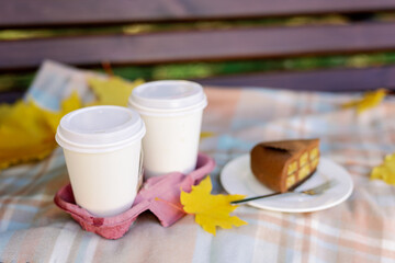 A snack in an autumn park on a bench with a blanket. Two cups of coffee and a piece of chocolate cake on a plate.